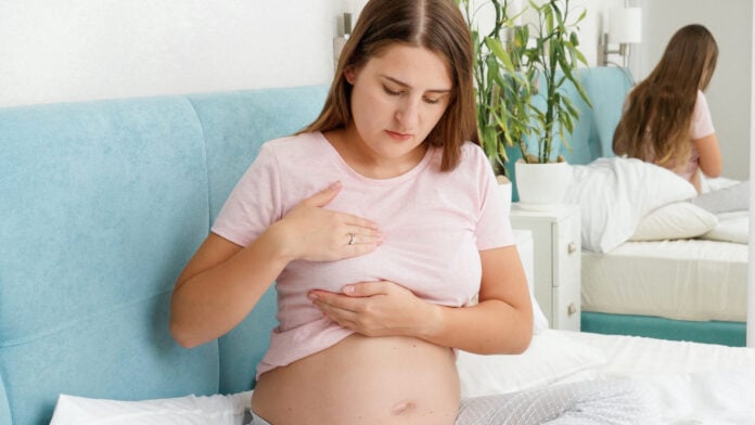 Schwangere Frau auf Bett hält schmerzende Brust und Bauch - typische Körperhaltung bei Mastitis-Symptomen