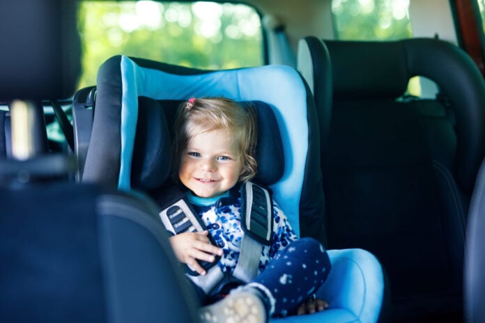 Fröhliches Kind mit blondem Haar sitzt sicher angeschnallt in einem blauen Kindersitz im Auto während einer Familienreise